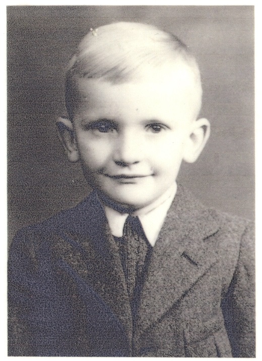 A black and white portrait of a young child with light-colored hair, wearing a collared shirt, tie, and a suit jacket. The child is looking directly at the camera with a subtle smile. The background is plain.