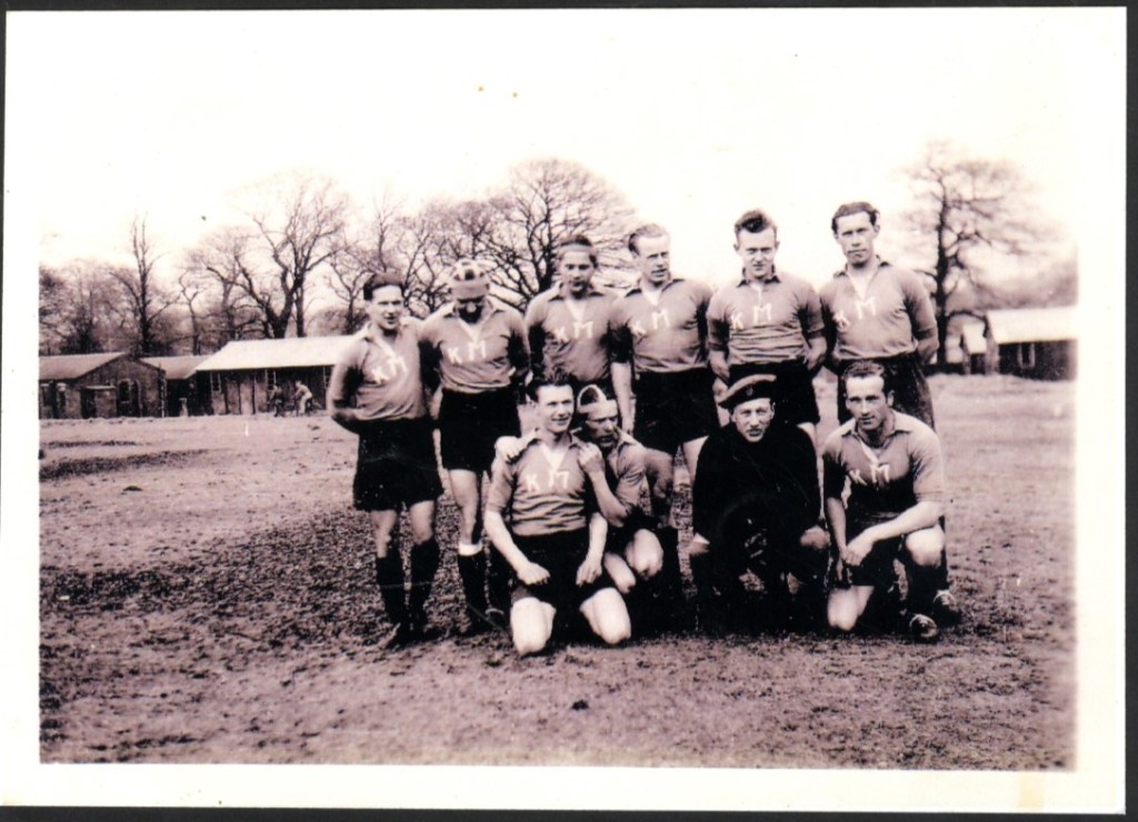 A black-and-white photo of a rugby team. Eleven men, dressed in matching jerseys and shorts, pose outside. Some are kneeling while others stand behind. Leafless trees and buildings are visible in the background. The mood appears friendly and relaxed.