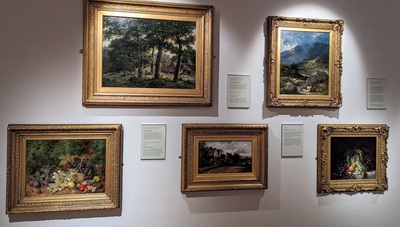 A museum wall displays five framed paintings. The paintings depict various landscapes and still lifes, including scenes of nature, mountains, fruit arrangements, and a village. Each painting is accompanied by a descriptive placard. The wall is lit to highlight the artwork.