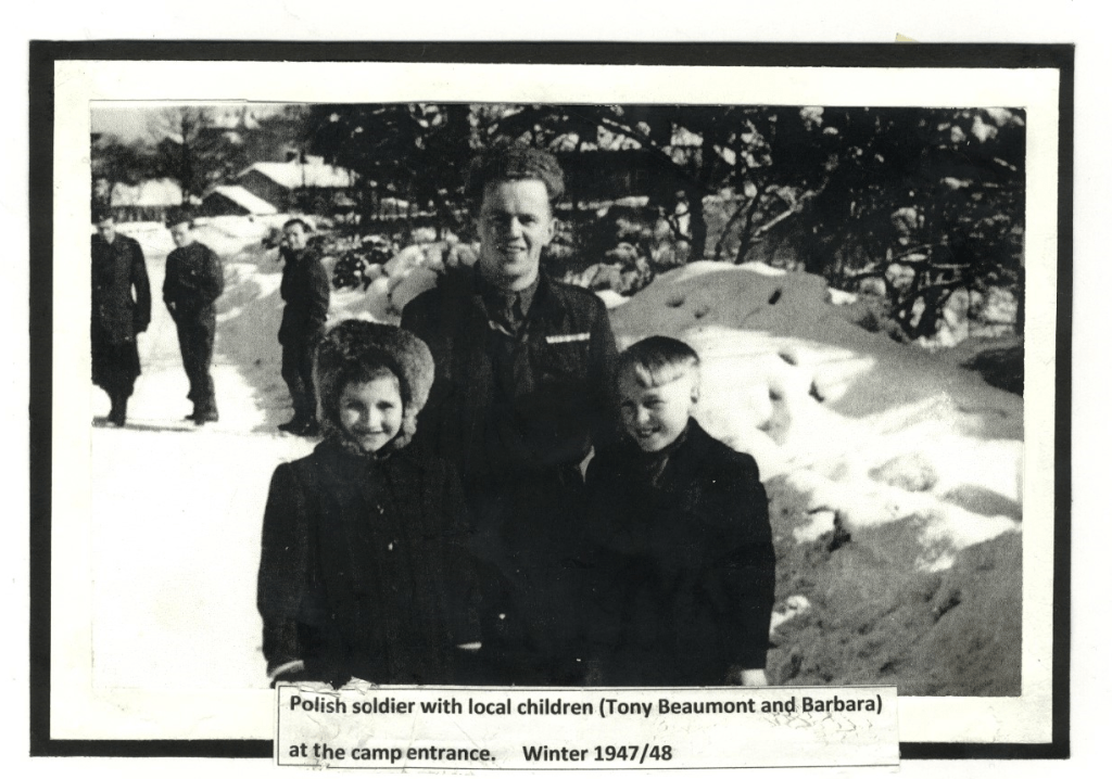 A black-and-white historical photograph depicts a smiling Polish soldier standing in snowy terrain, flanked by two local children. The scene is described as the camp entrance during winter 1947/48. In the blurred background, more people and snow-covered trees are visible.