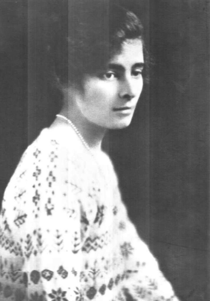 A black-and-white portrait of a woman from the early 20th century. She has short, wavy hair and is turned slightly to her left, gazing off to the side. She is wearing a patterned sweater with intricate designs and a string of pearls around her neck.
