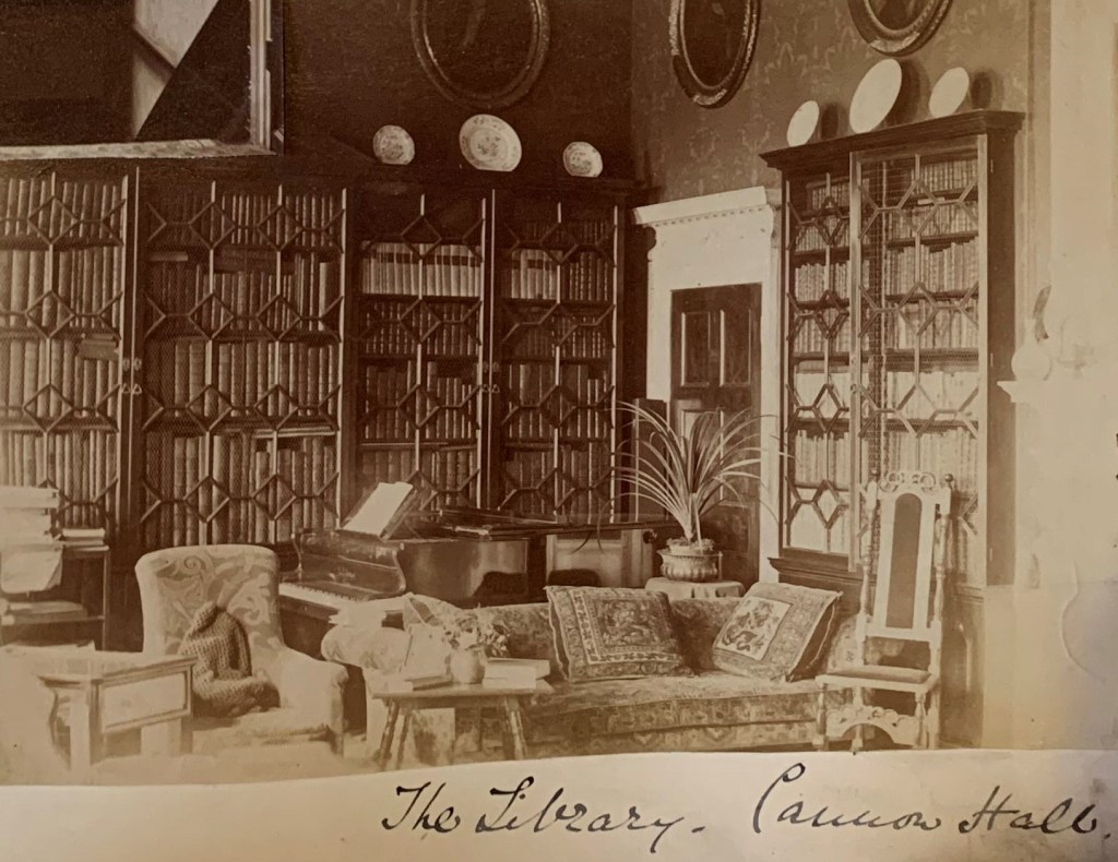 A vintage photo of a library room at Canons Hall. The room features tall bookshelves filled with books, a grand piano, a comfortable sofa, an armchair, and framed portraits on the walls. There is a handwritten note at the bottom that reads "The Library, Canons Hall.