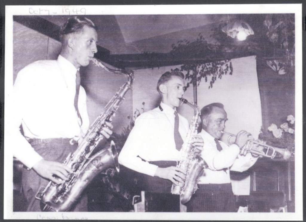 A black-and-white photograph taken in 1949 shows three men playing musical instruments in a band. The man on the left plays a saxophone, the man in the middle also plays a saxophone, and the man on the right plays a trumpet. All are wearing white shirts.