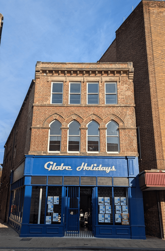A three-storey brick building with arched windows on the second floor and rectangular windows on the third. The ground floor has a blue storefront with the words "Globe Holidays" in white letters. Various posters are displayed in the lower windows.