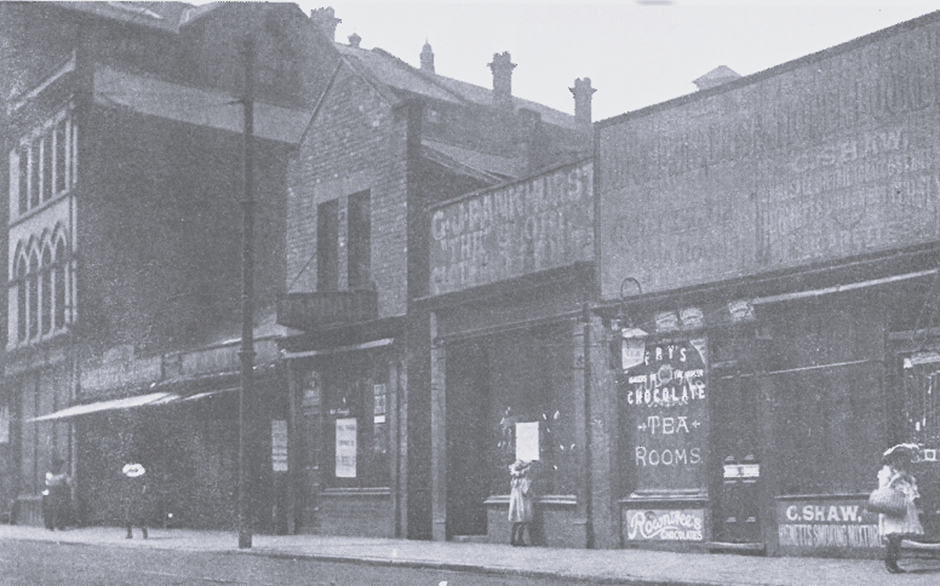 A vintage black-and-white photo shows a row of old brick buildings along Eldon Street. Signs in the windows display advertisements, and a few people are visible on the pavement.