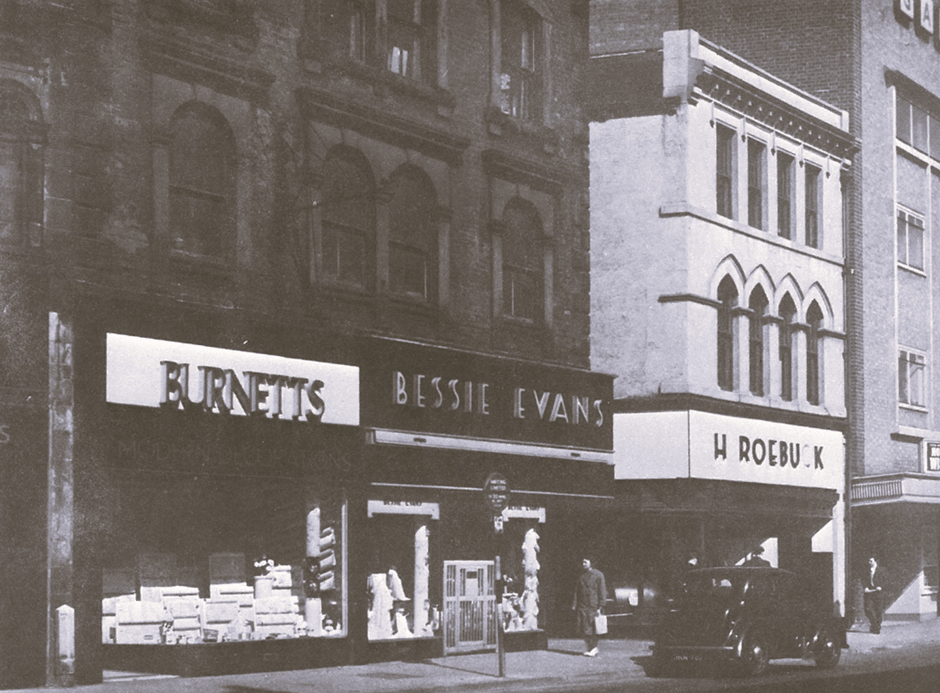 Black and white photograph of a row of shops, including Burnetts, Bessie Evans, and H Roebuck. The storefronts feature prominent signage, and merchandise is visible in the windows. A car is parked on the street in front of the buildings.