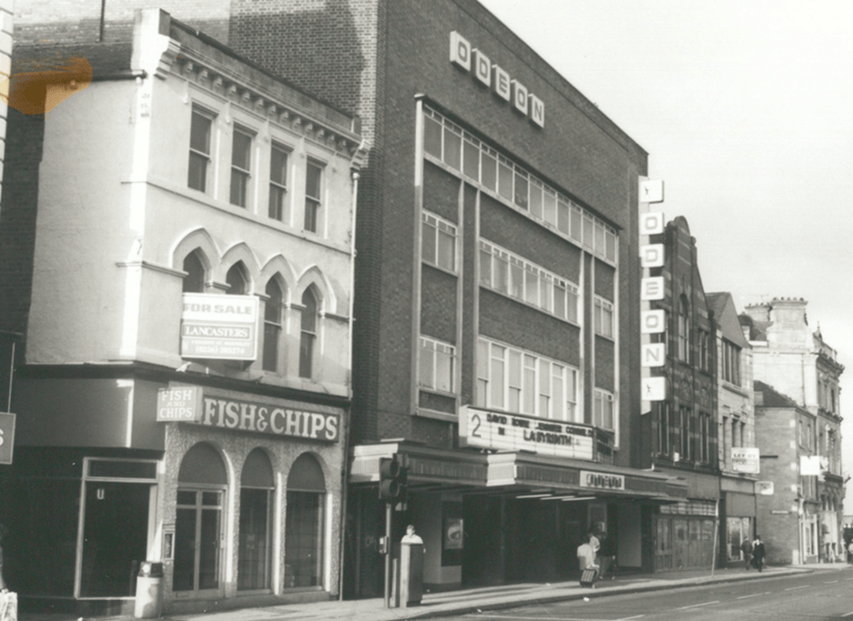 A black-and-white photo of Eldon Street with a fish and chip shop, a "For Sale" sign above it, and the Odeon cinema next door. The cinema read-o-graph shows movie titles. Other buildings and a few pedestrians are also visible.