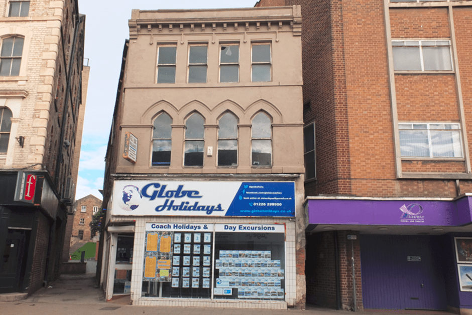 A street view of a travel agency named "Globe Holidays" located in a three-storey beige building with arched windows. Adjacent buildings are brick, with the one on the left slightly taller. A purple canopy is to the right. The sky is partly cloudy.