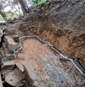 A small trench dug into a slope lined with stones and partially filled with mud and water. The trench is bordered by uneven dirt walls, and there's some greenery visible in the background. It appears the site is in a natural or wooded area.