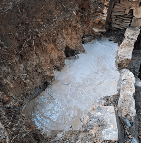 An excavation site reveals partially exposed stone foundations surrounded by soil. A layer of wet concrete covers part of the foundation, indicating recent construction or repair work. Logs and stones are piled in the corner of the site.
