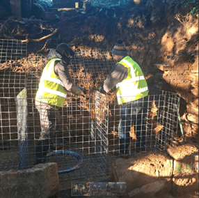 Two workers wearing high-visibility vests and beanies are constructing wire mesh cages outdoors. The area appears to be a construction site, and they are working on securing the mesh cages into the ground. The setting is sunny with shadows cast on the ground.