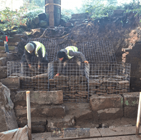 Two construction workers in safety vests and helmets are placing stones inside large metal wire cages on a rocky, partially excavated hillside. The cages are being filled to create a retaining wall structure. Vegetation and rock formations are visible in the background.