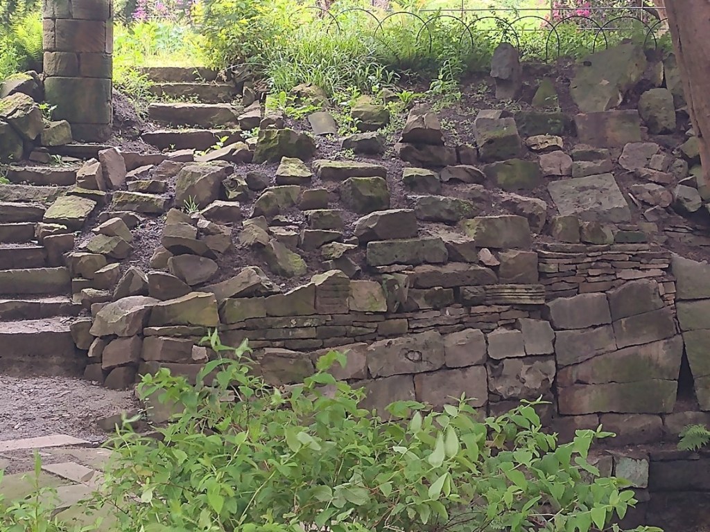 A stone pathway with moss-covered steps leads up to a rustic stone wall. The stones are unevenly stacked, and some are weathered. Green foliage is visible in the foreground, with more greenery and light filtering through trees in the background.