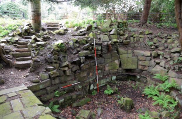 An old stone structure in the woods, partially restored with uneven bricks and steps leading upwards. Ferns and mosses grow among the stones, and a measuring rod stands vertically in the center, indicating the archaeological significance of the site.