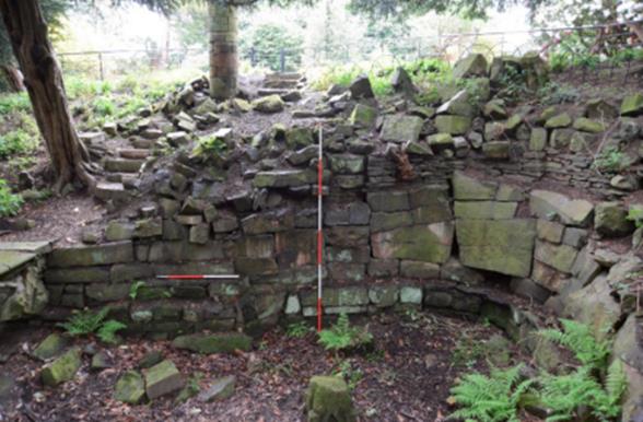 An archaeological excavation site featuring a partially excavated stone wall with large rocks. A measuring rod is placed vertically in the center, and a horizontal measuring scale is on the left side. The area is surrounded by trees and vegetation.