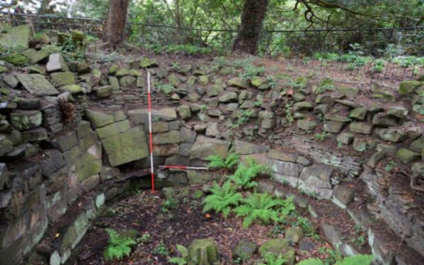 A circular stone structure, partially excavated and overgrown with ferns and moss, is nestled in a wooded area. Measuring rods are placed within the structure. The surrounding area is dense with trees and foliage, and an old iron fence can be seen in the background.