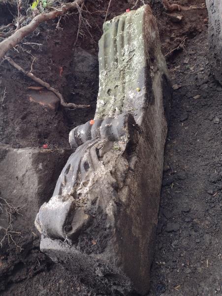 A large, ancient stone sculpture partially buried in dark soil. The sculpture appears to be intricately carved, displaying what might be parts of a figure or monument. Roots and rocks are visible around the sculpture, indicating it is part of an archaeological site.