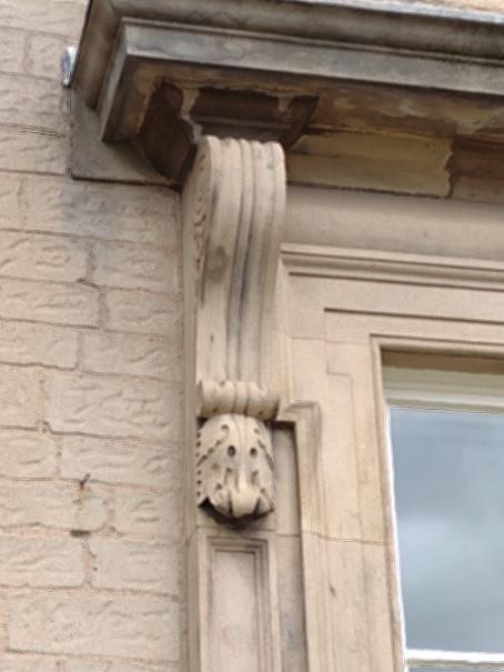 A close-up of a decorative architectural detail on a building. The stone carving features a scroll design with an ornate leaf and flower motif near the window frame. The surrounding stonework shows signs of aging and weathering.