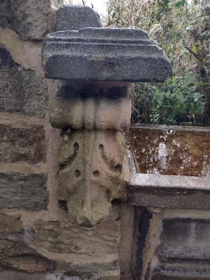 A stone wall with an ornate, weathered stone corbel supporting a small ledge. The corbel features carved decorative patterns and is partially covered with moss. The background shows greenery and another stone structure.