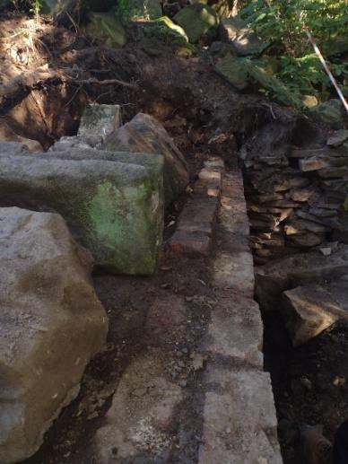 A partially excavated stone structure with a series of weathered, uneven steps leading down into the ground. The surrounding area is covered in soil and scattered rocks, with some greenery visible in the background.