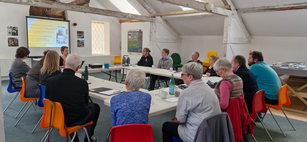 A group of eleven people wearing high-visibility vests and gloves stand in a green, wooded area. They are smiling at the camera, appearing to be part of a community cleanup or volunteer effort. The ground is covered in freshly cut vegetation.