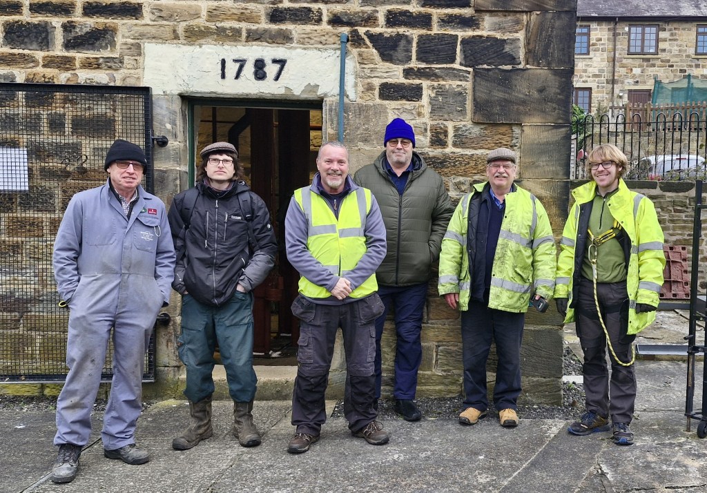 A group of eleven people wearing high-visibility vests and gloves stand in a green, wooded area. They are smiling at the camera, appearing to be part of a community cleanup or volunteer effort. The ground is covered in freshly cut vegetation.