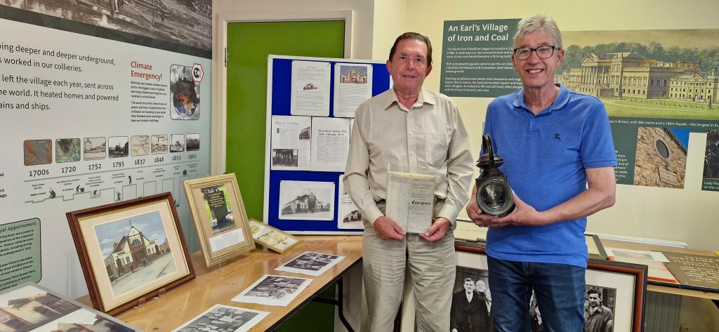 Two men stand indoors at an exhibition. The man on the left holds a book, and the man on the right holds a small trophy. Displays on the table include historical photos, documents, and memorabilia. Posters on the walls provide historical context of coal mining.
