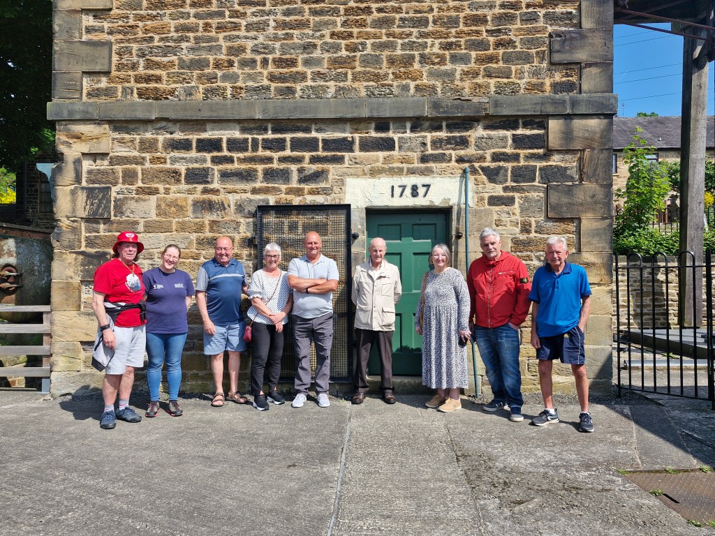 A group of nine people stand in front of a historic stone building, dated 1787 above the door. The individuals are casually dressed, with some wearing hats and sunglasses. The sun is shining brightly, casting shadows on the ground.