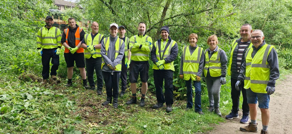 A group of eleven people wearing high-visibility vests and gloves stand in a green, wooded area. They are smiling at the camera, appearing to be part of a community cleanup or volunteer effort. The ground is covered in freshly cut vegetation.