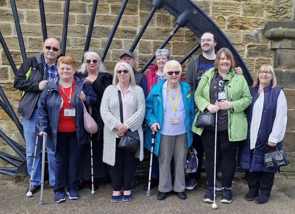 A group of ten people stands and smiles in front of a large, old-fashioned stone wall with an oversized wheel structure behind them. Some individuals hold white canes, suggesting that they may have visual impairments. The group is dressed in casual clothing.
