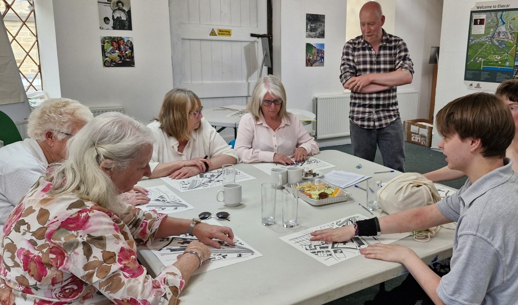 A group of six people sit around a table in a classroom-like setting, participating in an arts and crafts activity. They are leaning over printed papers. One person stands nearby observing. The table has mugs, glasses of water, and a plate of snacks on it.