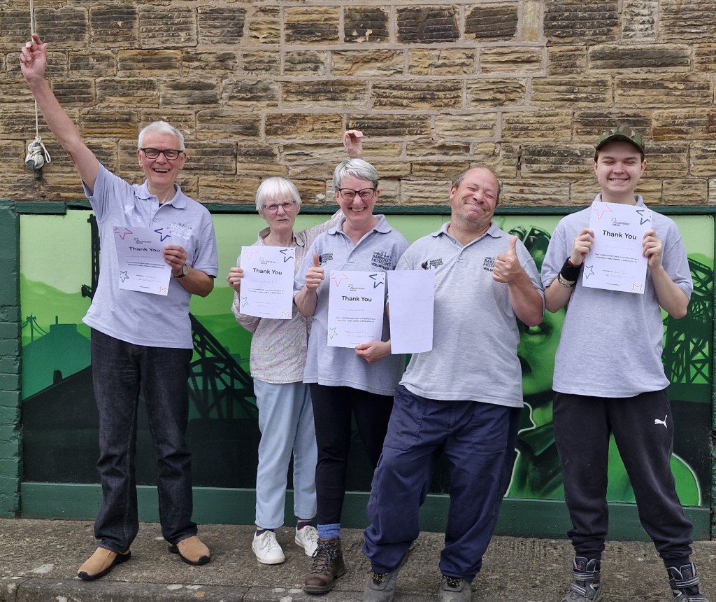 Five people smile and celebrate in front of a mural on a brick wall. They are holding thank you certificates and raising their hands victoriously. All are wearing matching gray shirts and dark pants. The background features a green mural with industrial elements.