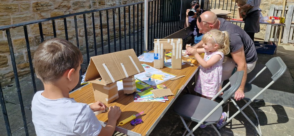 Children and an adult engage in a crafting activity at a long table outdoors. One boy assembles a structure from cardboard pieces, while a young girl focuses on her project with the help of the adult. Various crafting materials and tools are scattered on the table.