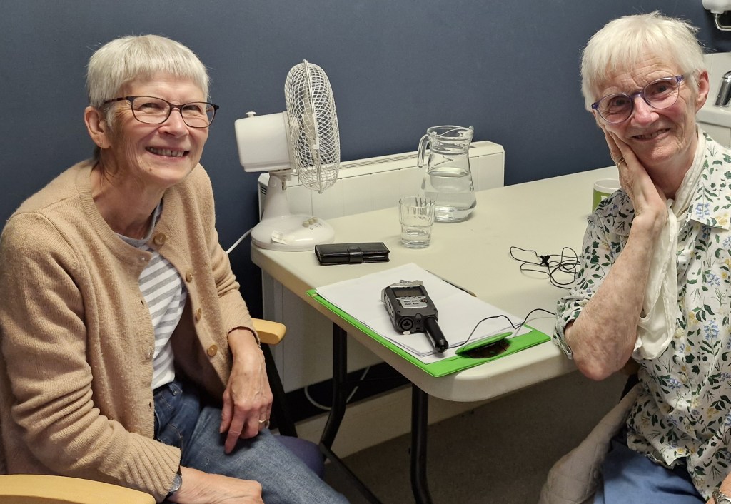 Two older women are seated at a table with a recording device, notebook, and glass of water. Both women are smiling at the camera; one wears a tan cardigan and striped shirt, the other a patterned blouse. A small fan and a lamp are visible on the blue wall behind them.