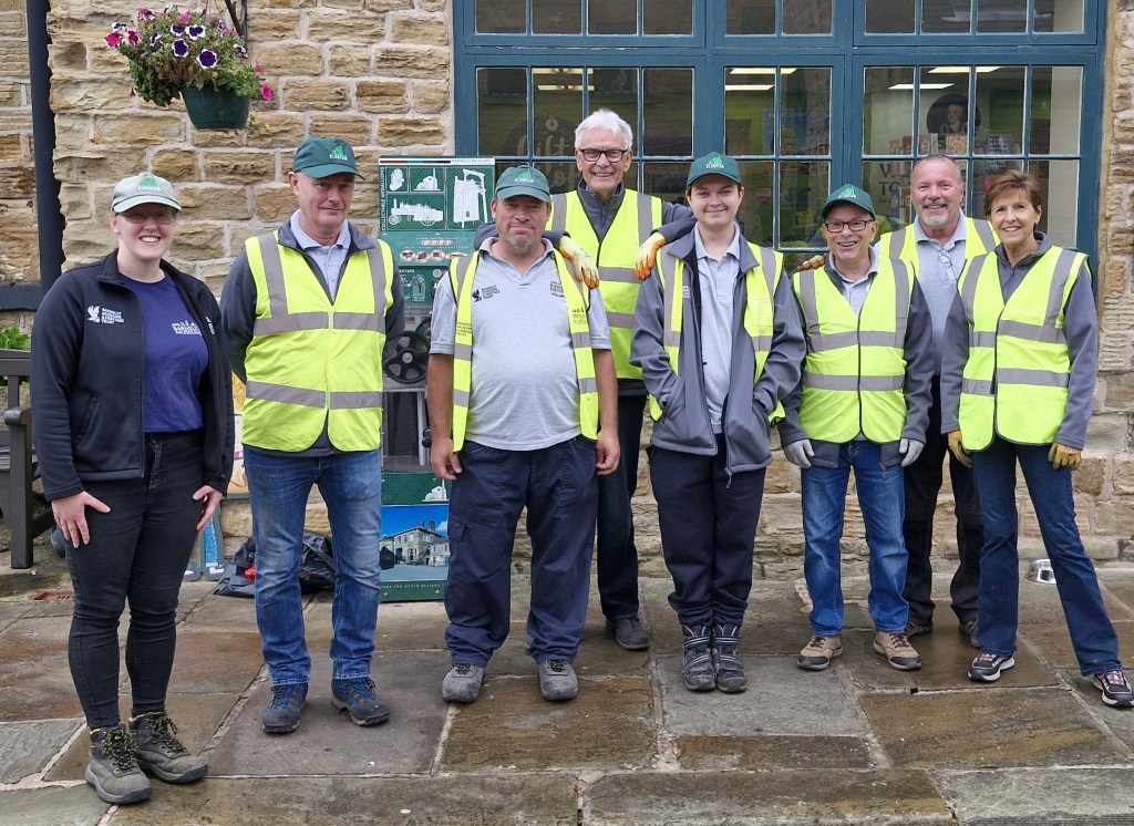 A group of eight people wearing high-visibility vests and casual clothing stand in front of a stone building with large windows. Some of them wear caps, and they all smile at the camera. Flowers in a hanging basket are visible on the left.