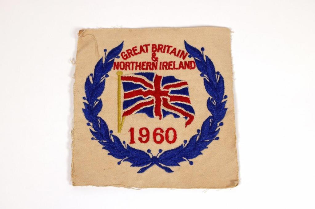 Black and white photo of a crowded stadium with spectators watching a track and field race. Several runners are visible on the track, with the crowd focused on the competition. The scene captures the excitement and anticipation of the event.