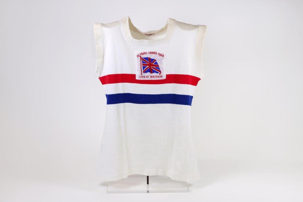 Black and white photo of a crowded stadium with spectators watching a track and field race. Several runners are visible on the track, with the crowd focused on the competition. The scene captures the excitement and anticipation of the event.