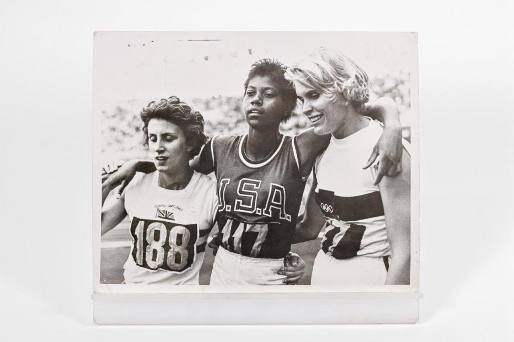Black and white photo of a crowded stadium with spectators watching a track and field race. Several runners are visible on the track, with the crowd focused on the competition. The scene captures the excitement and anticipation of the event.