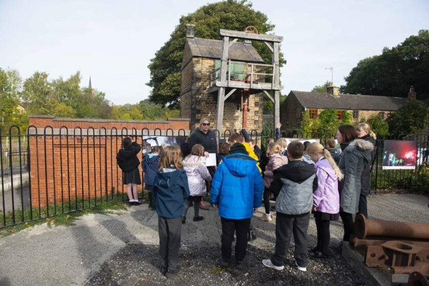 A group of children and a guide stand near an old stone building with an industrial structure on top, possibly a mine or mill. They are outdoors surrounded by trees and other buildings. A black railing and brick wall are also visible in the background.