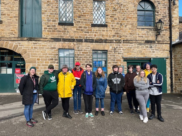 A group of twelve people, wearing casual winter clothing, stand smiling in front of an old brick building with green doors and window frames. The building has a rustic appearance, and the group looks happy to be together.