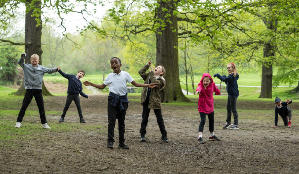 A group of children are playing energetically in a lush, green park. They are spread out with arms raised and wide smiles, enjoying the outdoors under tall trees with light green leaves. The kids are dressed casually in jackets, hoodies, and t-shirts.