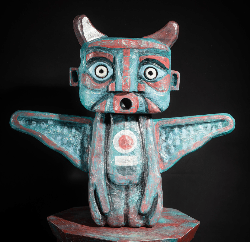 A colorful, wooden sculpture of a mythical creature with wings. The figure has large, round eyes, a gaping mouth, and protruding horns. The wings are wide and patterned. The sculpture is set against a black background, highlighting its vibrant hues.