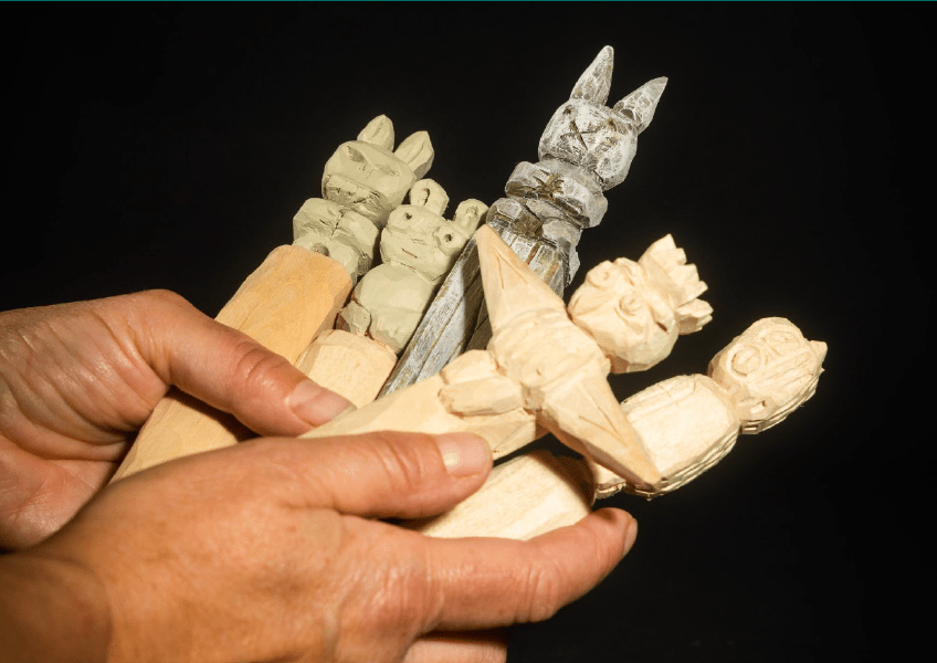 Two hands hold several partially carved wooden figures against a black background. The figures, all in various stages of completion, include simple animal shapes and human-like forms, showcasing different textures and details.