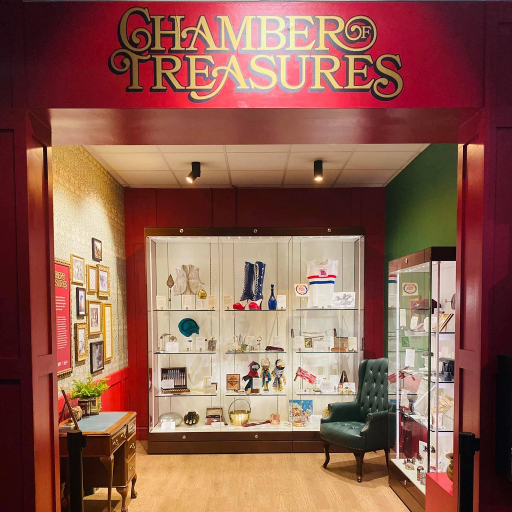 Black and white photo of a crowded stadium with spectators watching a track and field race. Several runners are visible on the track, with the crowd focused on the competition. The scene captures the excitement and anticipation of the event.