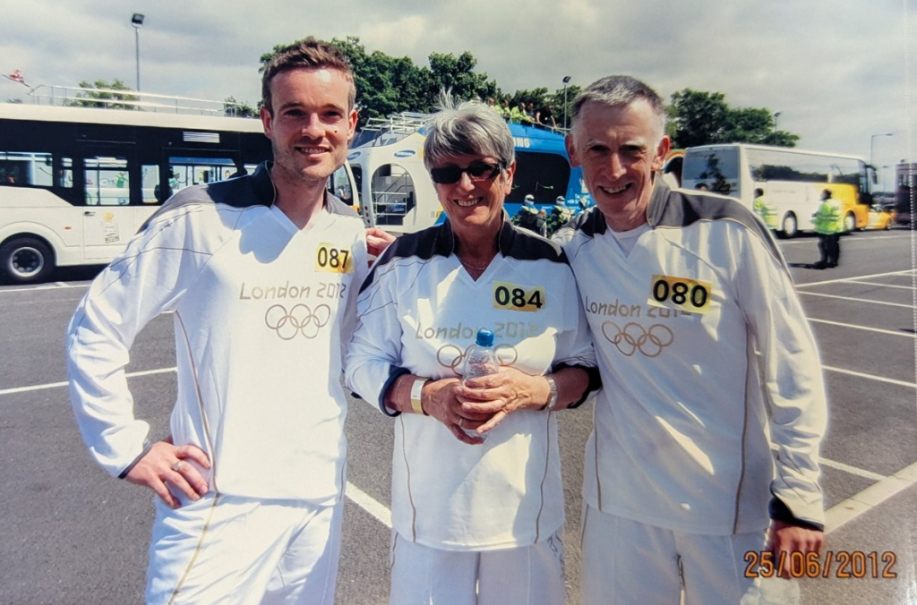 Black and white photo of a crowded stadium with spectators watching a track and field race. Several runners are visible on the track, with the crowd focused on the competition. The scene captures the excitement and anticipation of the event.