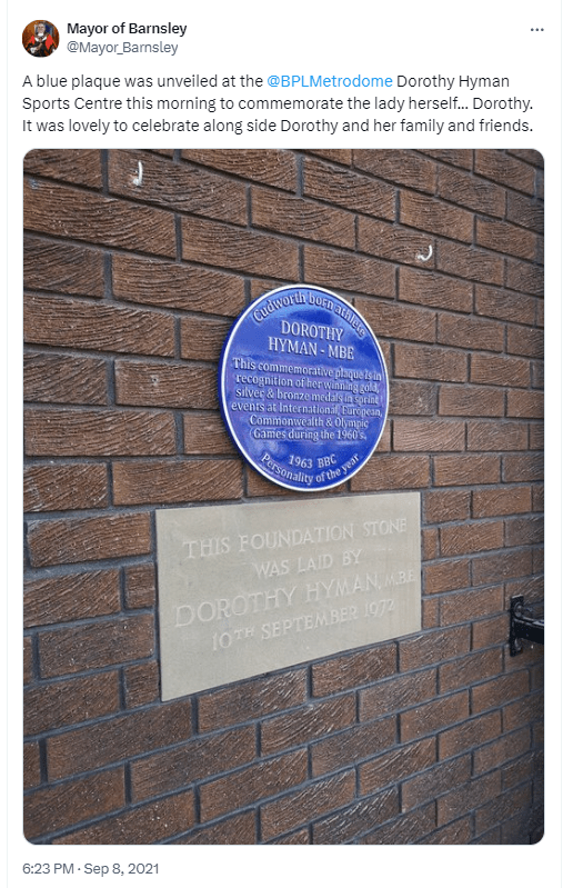 Black and white photo of a crowded stadium with spectators watching a track and field race. Several runners are visible on the track, with the crowd focused on the competition. The scene captures the excitement and anticipation of the event.