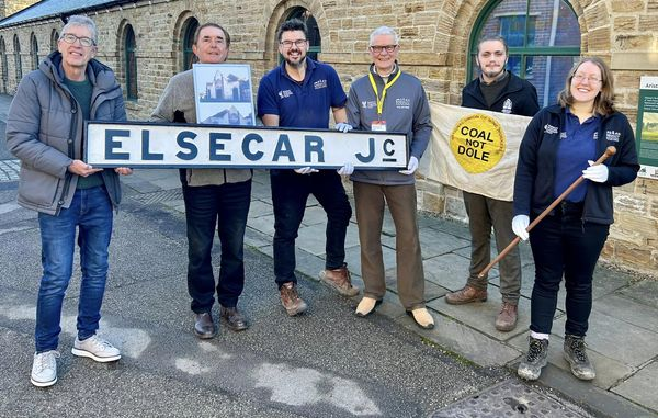 A group of six people stand outside a brick building, holding items including a large "Elsecar Jc" sign and a "Coal Not Dole" flag. They are all smiling at the camera and appear to be in good spirits.