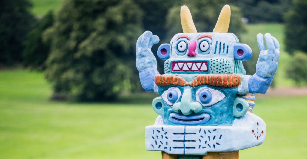 A colorful totem-like sculpture stands outdoors against a backdrop of green grass and trees. The sculpture features two stacked faces with horns and expressive features, each adorned with vibrant, intricate patterns. The scene is vibrant and artistic.