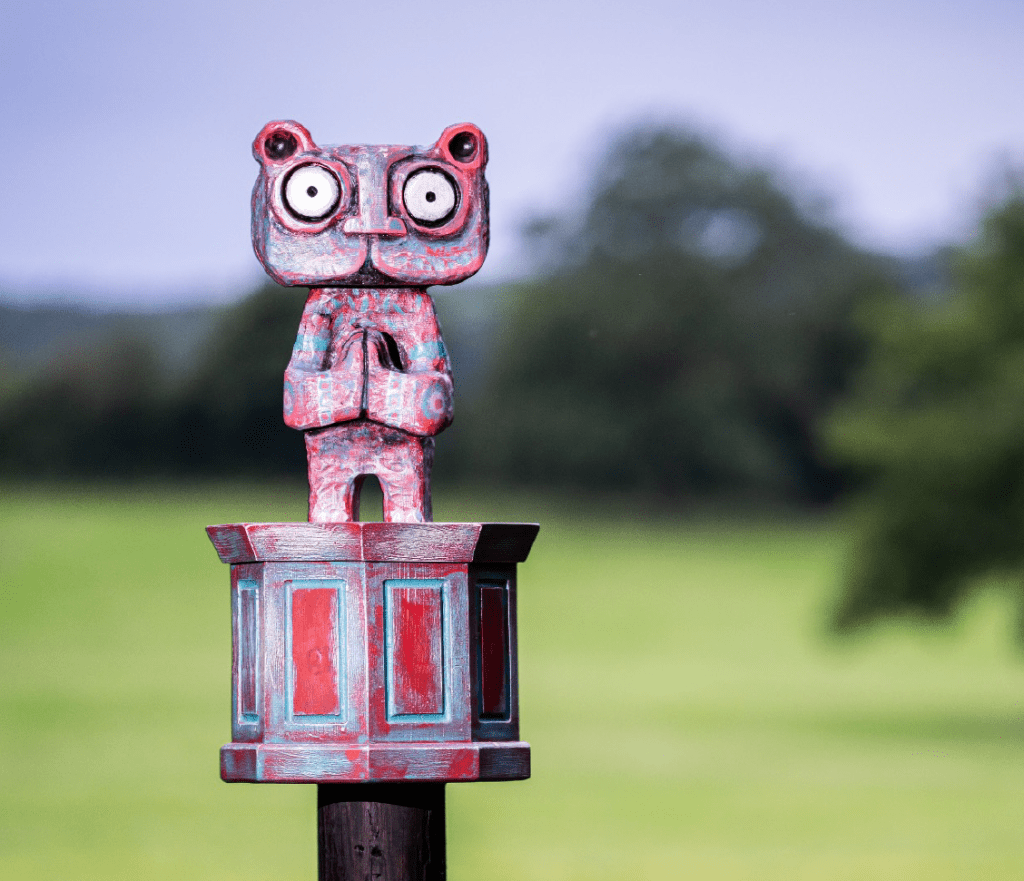 A colorful totem-like sculpture stands outdoors against a backdrop of green grass and trees. The sculpture features two stacked faces with horns and expressive features, each adorned with vibrant, intricate patterns. The scene is vibrant and artistic.