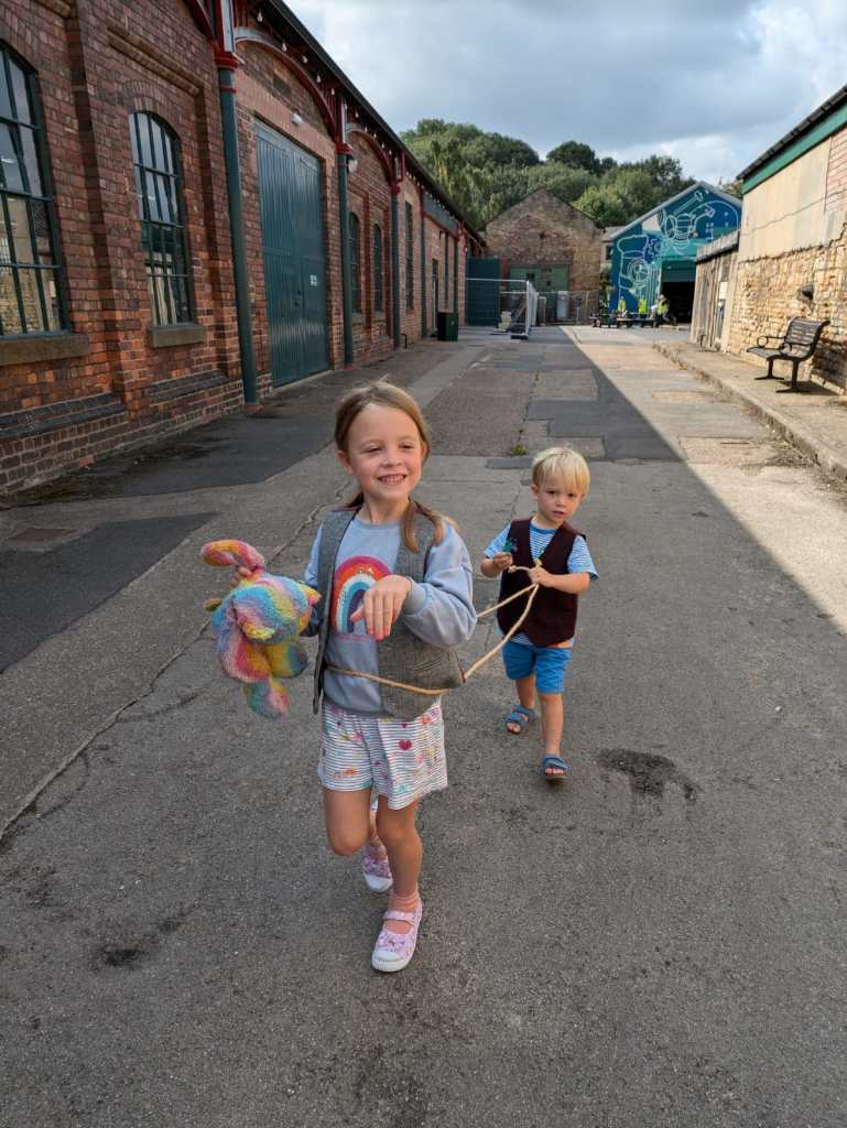 Two children are walking down a path between brick buildings. The older child, a girl, is holding a colorful stuffed animal and smiling. The younger child, a boy, is trailing behind her. Both look happy and are dressed casually. The sky is partly cloudy.